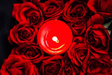 Parlak kırmızı güller zarif bir şekilde parlayan bir mumu çevreleyip romantik bir ortam yaratıyor..