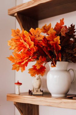 Beyaz bir sürahi, sonbaharın özünü yakalayan parlak turuncu yaprakları ahşap bir rafta sergiliyor. Sıcak renkler sıcak bir atmosfer yaratır..