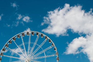 Güneşli bir öğleden sonra, tüylü beyaz bulutlarla kaplı parlak mavi bir gökyüzüne karşı büyük bir dönme dolap dik durur..