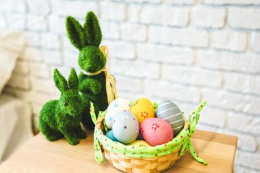 Parlak renkli Paskalya yumurtaları, ahşap bir yüzey üzerinde yosundan yapılmış iki yeşil dekoratif tavşanın yanında örülmüş bir sepetin içinde düzenlenir..