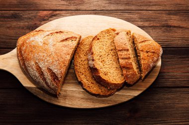 Sıcak taze pişmiş kahverengi ekmek ahşap bir kesim tahtasının üzerinde sergileniyor. Birkaç dilim kesilmiş ve düzgünce düzenlenmiş, dokusu ve rengi sergileniyor..