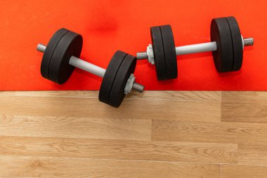 İki dambıl, ahşap bir spor salonunun zemininde parlak kırmızı bir paspasın üzerinde sabah antrenmanına hazır bekliyor..