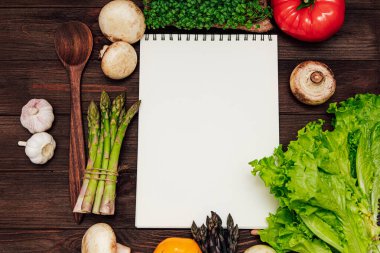 Taze sebzelerden oluşan parlak çeşitlilik, ahşap bir yüzey üzerindeki boş bir not defterinin etrafını sarar..