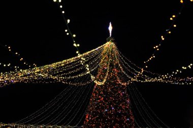 Gece karanlığında parıldayan süslemelerle çevrili büyük bir Noel ağacında parıldayan renkli ışıklar Noel ruhunu kutluyor..