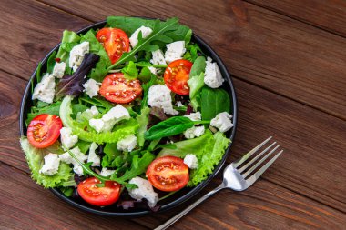 Karışık sebzeli, dilimlenmiş domatesli ve rendelenmiş peynirli canlı bir salata yanında çatalla kırsal ahşap bir masada sunuluyor..