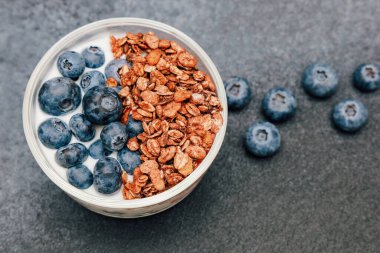 Yoğurtla dolu besleyici bir kahvaltı kasesi. Üzeri granola ve koyu bir tezgahta taze yaban mersini..