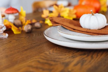 Renkli sonbahar yaprakları ve dekoratif eşyalar arasında güzel düzenlenmiş bir yemek masasında beyaz tabaklar turuncu peçeteler ve küçük balkabakları sergileniyor. Çekici mevsimsel bir toplantının ipuçları.