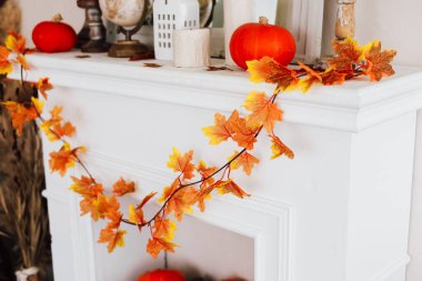 Mevsimlik dekor parlak turuncu balkabaklarıyla süslenmiş beyaz bir şömine rafı ve renkli sonbahar yapraklarından bir çelenk içerir. Sıcak davetkar düzenleme sonbahar şenlikleri için rahat bir ortam yaratıyor..