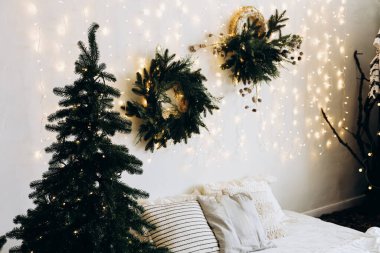 Sıcak bir kış yatak odasında küçük bir Noel ağacı ve duvarda güzel düzenlenmiş çelenkler bulunur. Telli ışıklar büyüleyici bir atmosfer yaratıyor. Tatil sezonunun şenlikli dekorunu geliştiriyor..