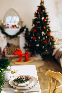 Güzel düzenlenmiş bir yemek masasında çam dalları ve baston şekerler gibi dekoratif elementler bulunur. Arka planda süslerle süslenmiş bir Noel ağacı samimi odaya neşe katar..