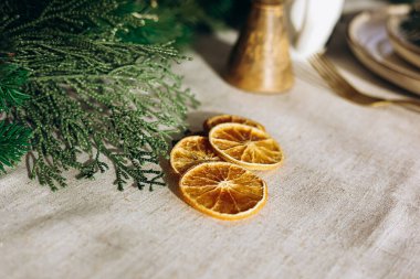 Kurutulmuş portakal dilimleri, Noel 'i dekoratif yeşilliklerle çevrili yumuşak bir masa örtüsünde düzenler. Ortam, sıcak ve davetkar atmosferi geliştiren bir tatil toplantısını öneriyor..