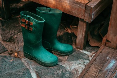 Yeşil lastik çizmeler, bahçedeki ahşap bankların yanında, taş bir patikada duruyor. Sahne sakin görünüyor. Açık hava macerası ya da bahçe işleri. Yumuşak ışık çizmeleri ve çevreyi vurgular.