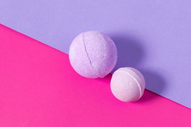 İki yuvarlak banyo bombası biri mor diğeri ise parlak pembe ve mor bir arka planda duruyor. Renkler ortaya çıkar ve rahatlamak için ideal neşeli bir atmosfer yaratır..