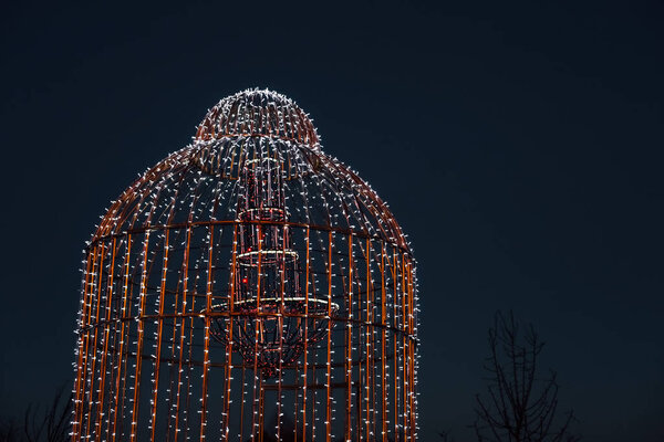 Красивая клетка, украшенная яркими огнями, стоит на фоне тёмного неба. Он создает завораживающую атмосферу, идеально подходящую для вечерних торжеств и банкетов во время праздничного сезона.