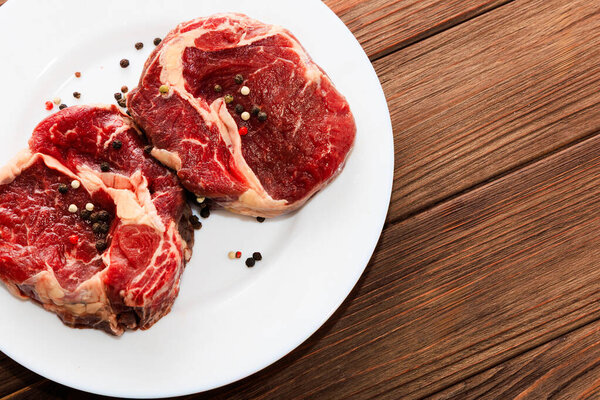 Two ribeye steaks are placed on a white plate sprinkled with assorted peppercorns. The steaks are raw and show marbled fat. The wooden table creates a rustic backdrop for the scene.