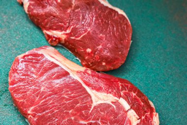 Two raw beef steaks are placed on a green cutting board in a kitchen. The meat displays rich red colors and marbling ready for preparation and cooking. They appear fresh and high quality.
