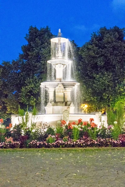 Beleuchteter Dreistufiger Brunnen Umgeben Von Blumen Unter Dunklem Nachthimmel — Stockfoto