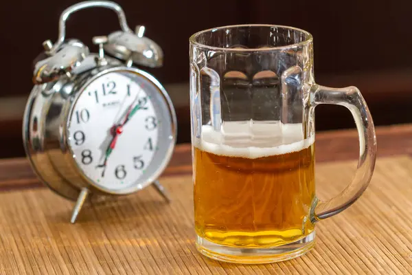 ビールと古い銀の目覚まし時計の半分のフルガラスマグ ストック画像