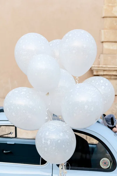 Various white balloons tied to white automobile with sand colored stone wall from building in background