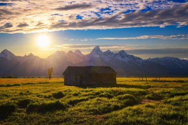 Summer sunset over a historic farm shed at Mormon Row in Grand Teton National Park, with snowcapped Teton Mountain Range in the background.