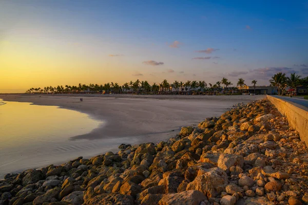 Sunset above Arabian Sea and a beach in Salalah, Oman, with palm trees, hotel resorts and rocky shoreline.