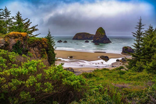 Coastal landscape at Samuel H. Boardman State Scenic Corridor, featuring sandy beach, rocky sea stacks, and lush greenery under a cloudy sky
