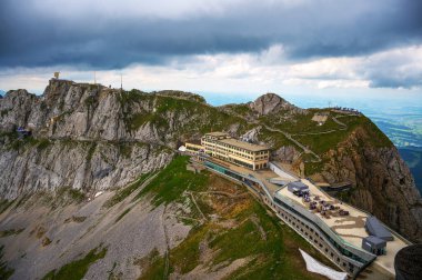 Aerial view of Mount Pilatus in Switzerland, showcasing its hotel resorts, rocky cliffs, and green terrain under a cloudy sky. clipart