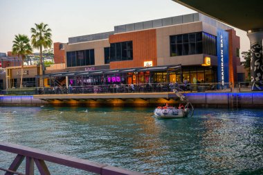 Manama, Bahreyn - 27 Aralık 2023: Bahreyn, Manama 'daki Bahreyn Körfezi' nde akşam manzarası, bir rıhtım restoranı, modern binalar ve su üzerinde yolcuları olan bir tekne.