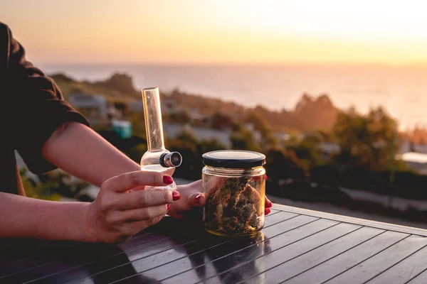 大きなマリファナの芽とボンボン 雑草の水パイプ と美しい夕日の空と海を持つガラス瓶を保持赤い爪を持つ手 — ストック写真
