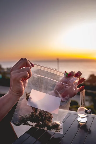 大きなマリファナの芽 とコピースペース ボング 雑草水パイプ と美しい夕日の空と海を持つビニール袋を保持赤い爪を持つ手 — ストック写真