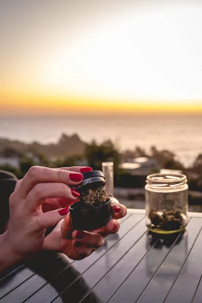 マリファナのグラインダーに雑草の芽を入れて赤い爪を持つ手 美しい夕日の空と海とマリファナの芽と水パイプ を持つガラス瓶 — ストック写真