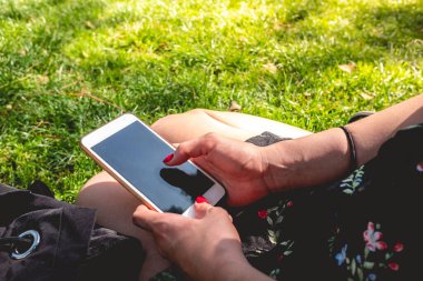 Kırmızı tırnaklı kız elleri, güneşli bir günde parkta çimlerin üzerinde siyah ekranlı beyaz bir akıllı telefon tutuyor.