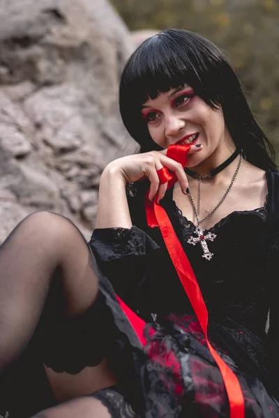 太陽の下で大きな岩の間の木のベンチに 赤いリボン で座って遊んで黒のドレスを着た若い セクシーで吸血鬼のヒスパニックゴスの女の子 — ストック写真