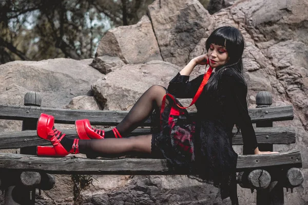 黒のドレスと赤いプラットフォームの靴を着た若い セクシーで吸血鬼のヒスパニックゴスの女の子は 日光の下で大きな岩の間の木のベンチで 赤いリボン で座って遊んでいます — ストック写真