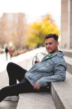 Güneş gözlüklü, yeşil tişörtlü, kot ceketli ve siyah kotlu genç sarışın adam öğleden sonra şehirdeki bir parkta taş merdivenlerde oturuyor.