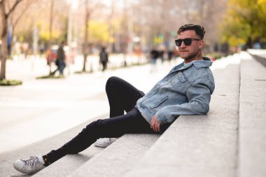 Güneş gözlüklü, yeşil tişörtlü, kot ceketli ve siyah kotlu genç sarışın adam öğleden sonra şehirdeki bir parkta taş merdivenlerde oturuyor.