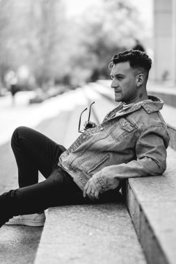 Güneş gözlüklü, kot ceketli ve siyah kotlu genç çekici adam öğleden sonra şehirdeki bir parkta taş merdivenlerde oturuyor.)