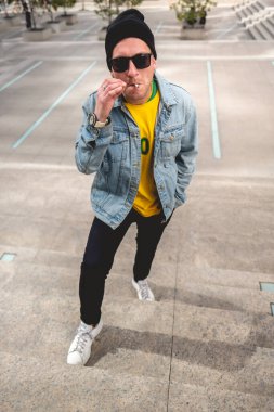 Güneş gözlüklü, sarı tişörtlü, kot ceketli, siyah yün şapkalı ve siyah kotlu genç adam öğleden sonra şehirdeki bir parkta taş merdivenlerde durup sigara içiyor.