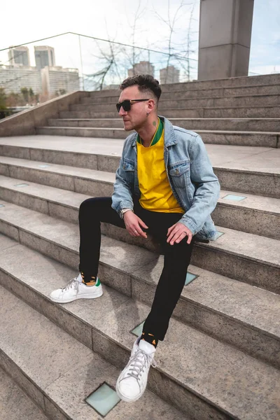 Young blonde model guy with 4-leaf clover and sunglasses, yellow t-shirt, denim jacket and black jeans sitting on stone stairs in a park in the city in the afternoon