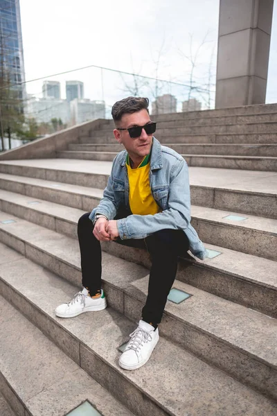 Young blonde model guy with sunglasses, yellow t-shirt, denim jacket and black jeans sitting on stone stairs in a park in the city in the afternoon