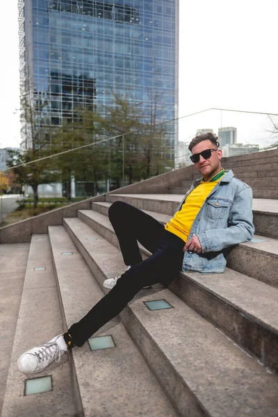 若いですブロンドモデル男とともにサングラス 黄色のTシャツ デニムジャケットと黒ジーンズに座って石の階段で街の公園で午後 — ストック写真