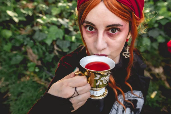 赤い毛のスカーフと暗い服を着た若い赤い髪の女の子は 公園で古い世紀のティーカップとソーサーで赤い飲み物 を飲む — ストック写真