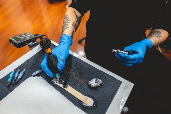 Hands of a tattoo artist holding a tattoo machine charging black ink, and work table with needles, plastic ink containers, alcohol and napkins