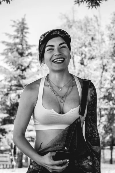Happy, sexy and tattooed young girl with cap, denim dungarees and white crop top smiling, laughing and holding a phone in the park in a sunny day (in black and white)