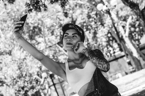 Happy, sexy and tattooed young girl with cap, denim dungarees and white crop top smiling, laughing and making a video call on the phone in the park (in black and white)
