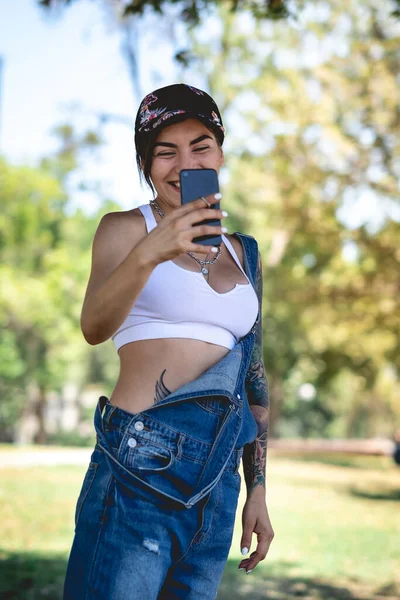 Happy, sexy and tattooed young girl with cap, denim dungarees and white crop top smiling, laughing and holding a phone in the park in a sunny day