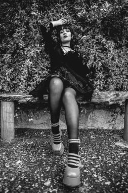 Siyah elbiseli ve platformlu genç ve sıska İspanyol gotik kız. Kırsal bir ahşap bankta oturan ayakkabılar. Ormandaki beyaz çiçeklerin yanında.)