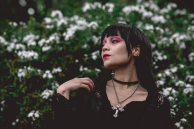 Siyah elbiseli ve kırmızı makyajlı genç ve sıska İspanyol gotik kız ormanda beyaz çiçeklerin yanında poz veriyor.