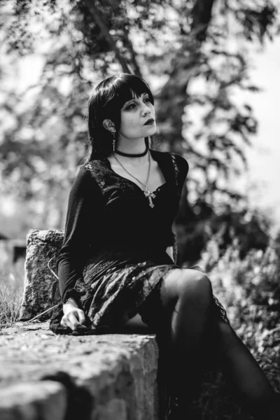 Young and skinny hispanic goth girl with black dress and red platforms shoes seated in a little stones wall in the forest (in black and white)