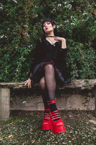 若いですとスキニーHispanicゴス女の子とともに黒のドレスと赤のプラットフォームの靴で座っています素朴な木のベンチ森の中の白い花の横に — ストック写真